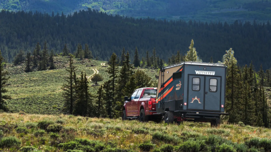 A red pickup truck towing a Winnebago HIKE 100 in a forest setting